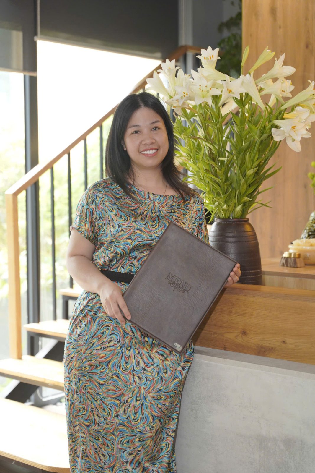 Chị Vân Anh cầm cuốn menu bên quầy gỗ và bình hoa loa kèn trắng, nụ cười của một người đã đứng trước mặt khách hơn hai mươi hai năm.