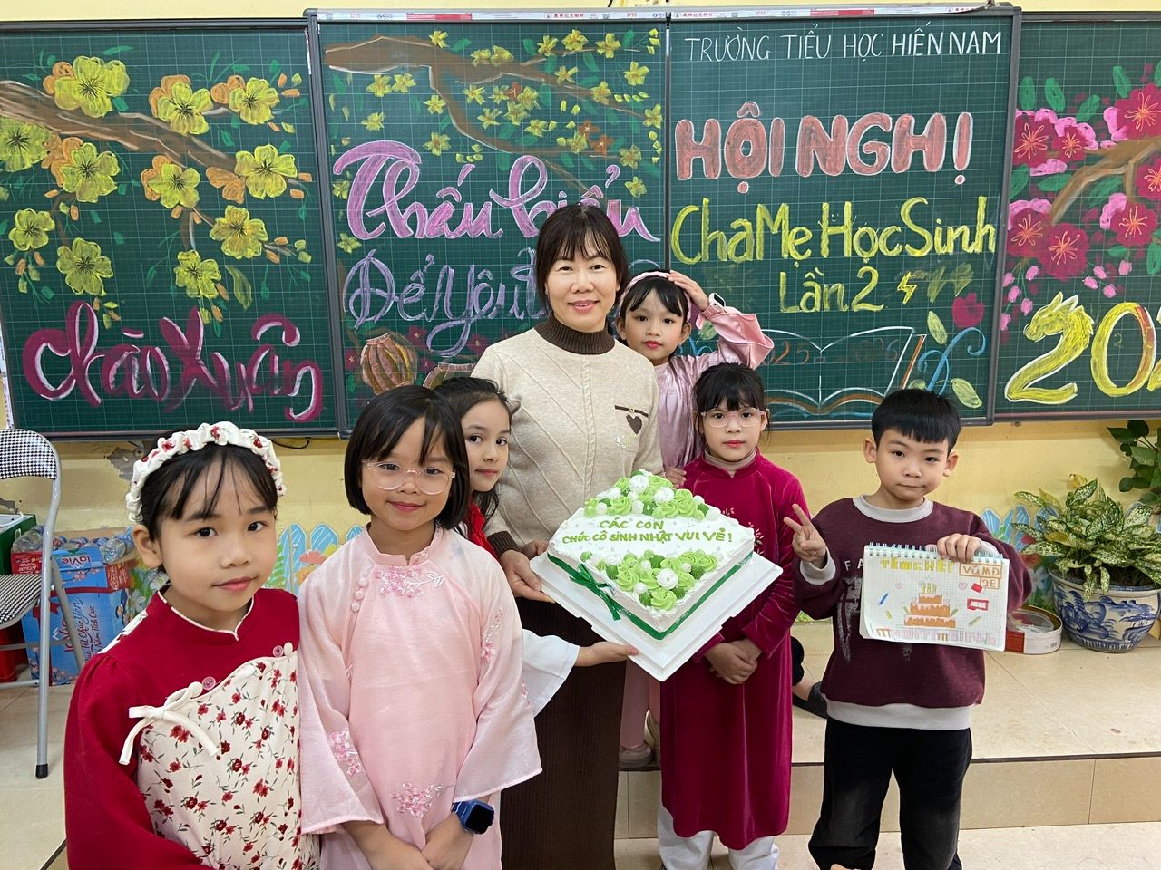 Ngày sinh nhật cô Thuỷ, các con trong lớp bưng chiếc bánh ghi dòng chữ "Các con chúc cô sinh nhật vui vẻ", một cậu bé giơ tấm thiệp tự vẽ.