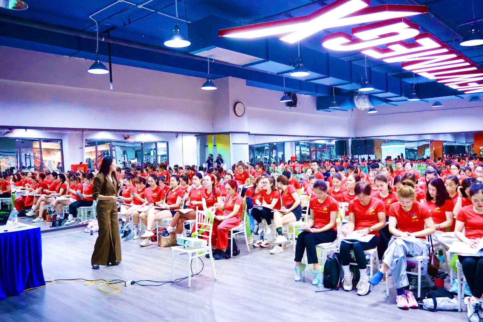 Chị Thơ đứng lớp giữa một phòng tập kín chỗ ngồi, những huấn luyện viên mặc áo đỏ đồng phục cúi đầu ghi chép.