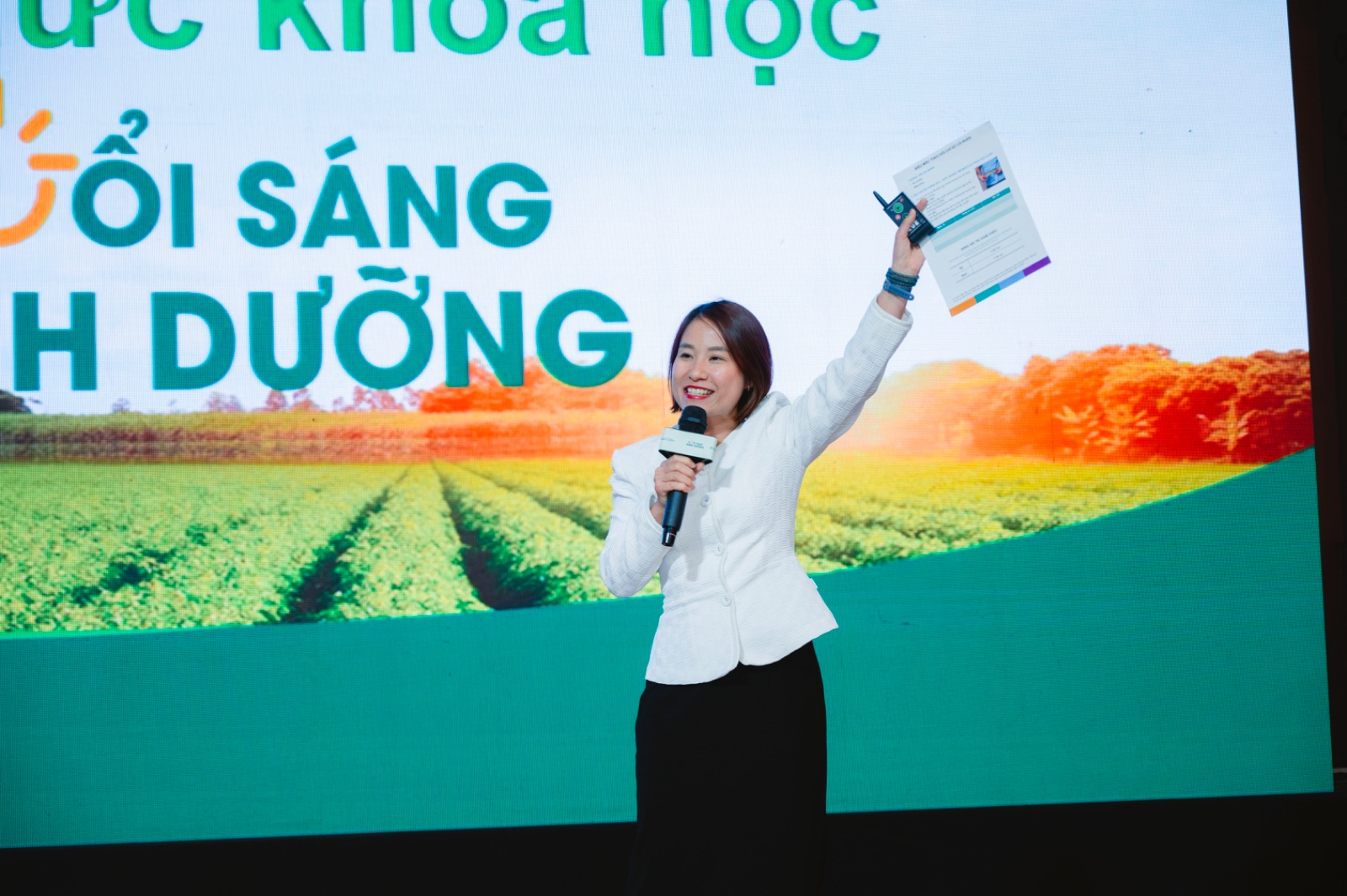 Chị Hằng cầm micro đứng giảng trước màn hình chủ đề dinh dưỡng, tay giơ cao tờ tài liệu cho cả hội trường cùng nhìn.