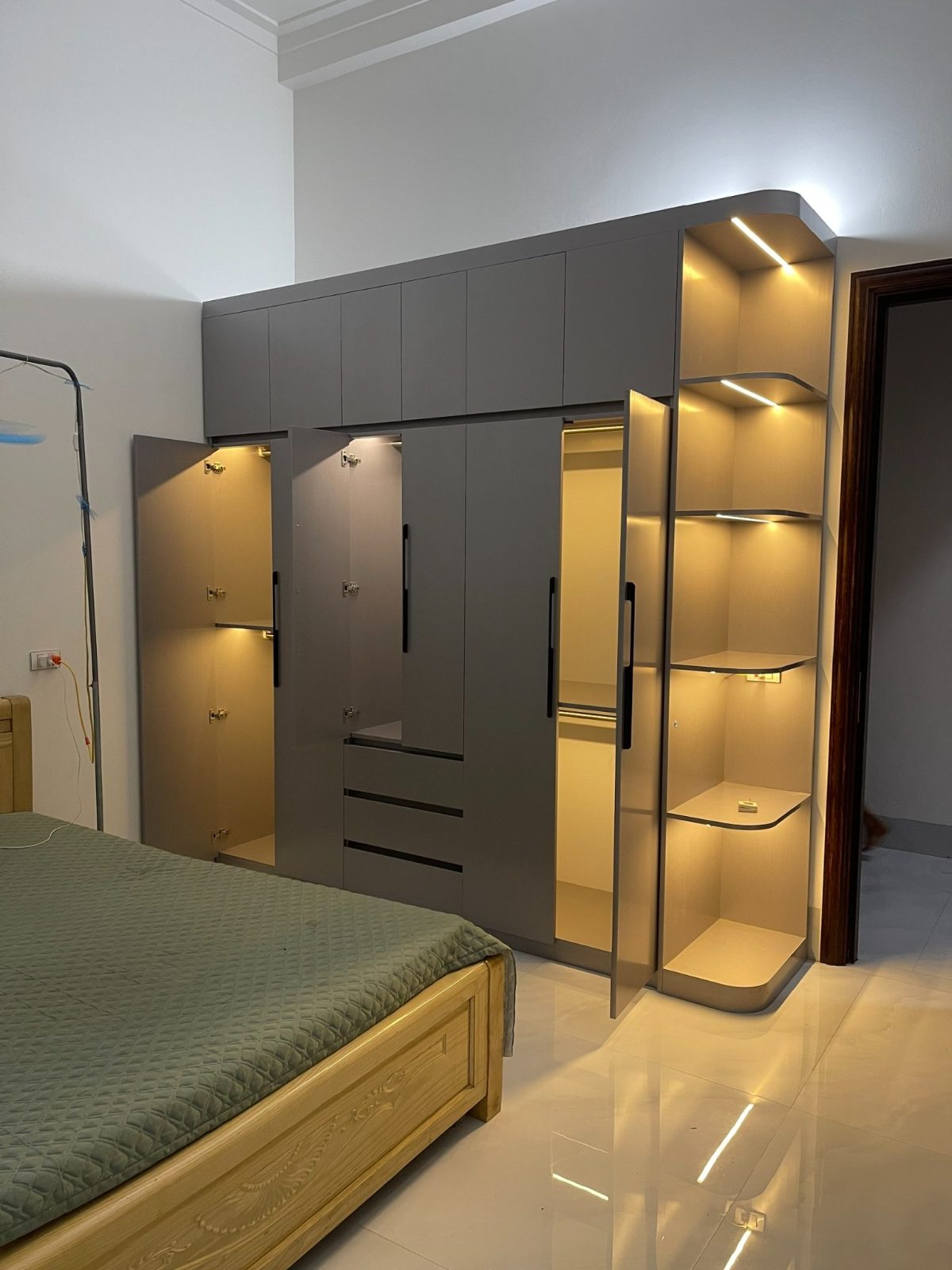 Bộ tủ áo kịch trần màu ghi do xưởng của anh Đông hoàn thiện: cánh mở lộ khoang treo, ngăn kéo và dãy kệ góc bo tròn có đèn LED, đúng tinh thần bền trước, tiện sau, đẹp trọn của anh.