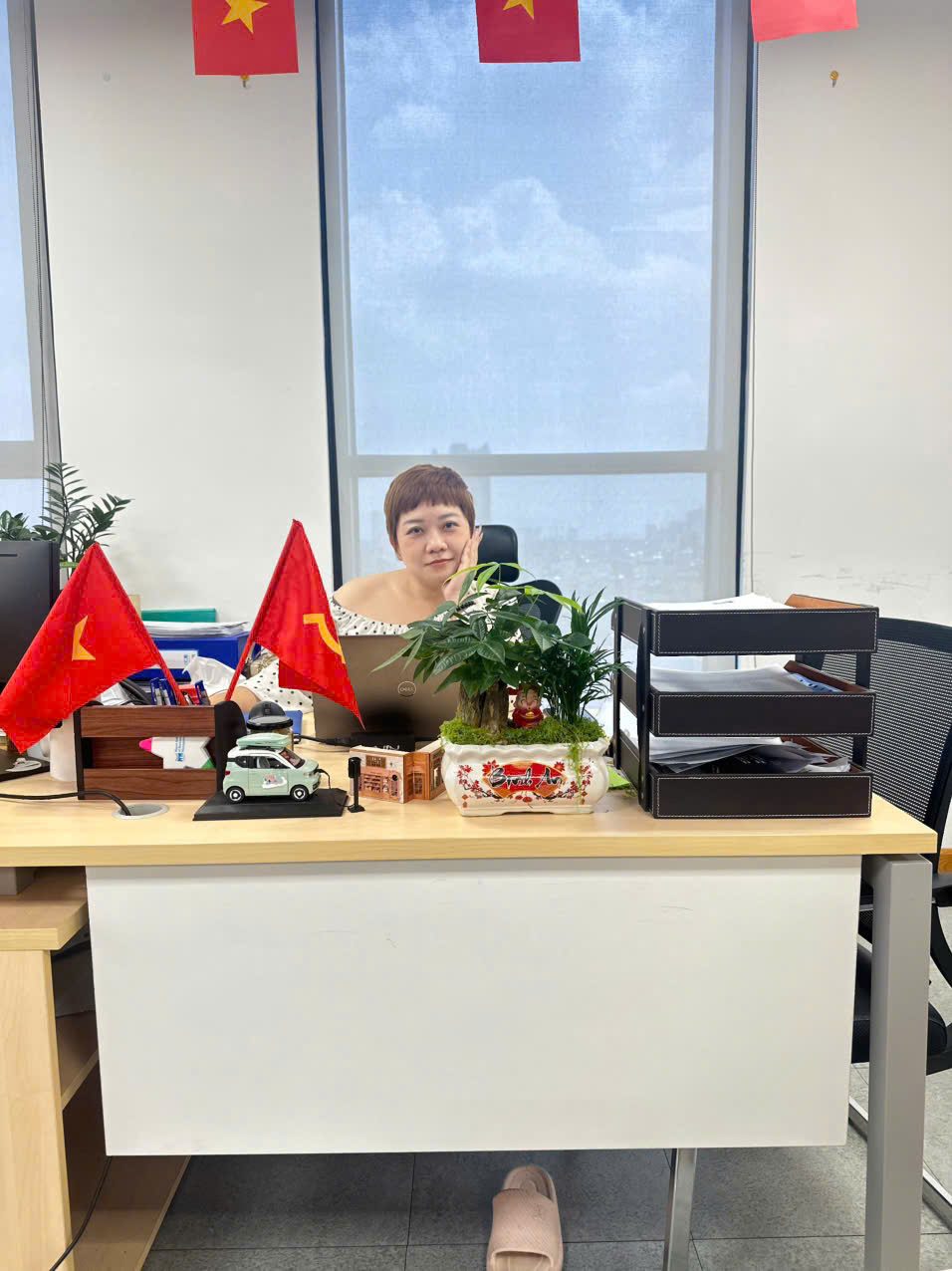 Chị Ngọc Saphia bên bàn làm việc, giữa laptop, khay hồ sơ và hai lá cờ đỏ sao vàng, nơi những bản kế hoạch truyền thông và kịch bản sự kiện được viết từ trang trắng