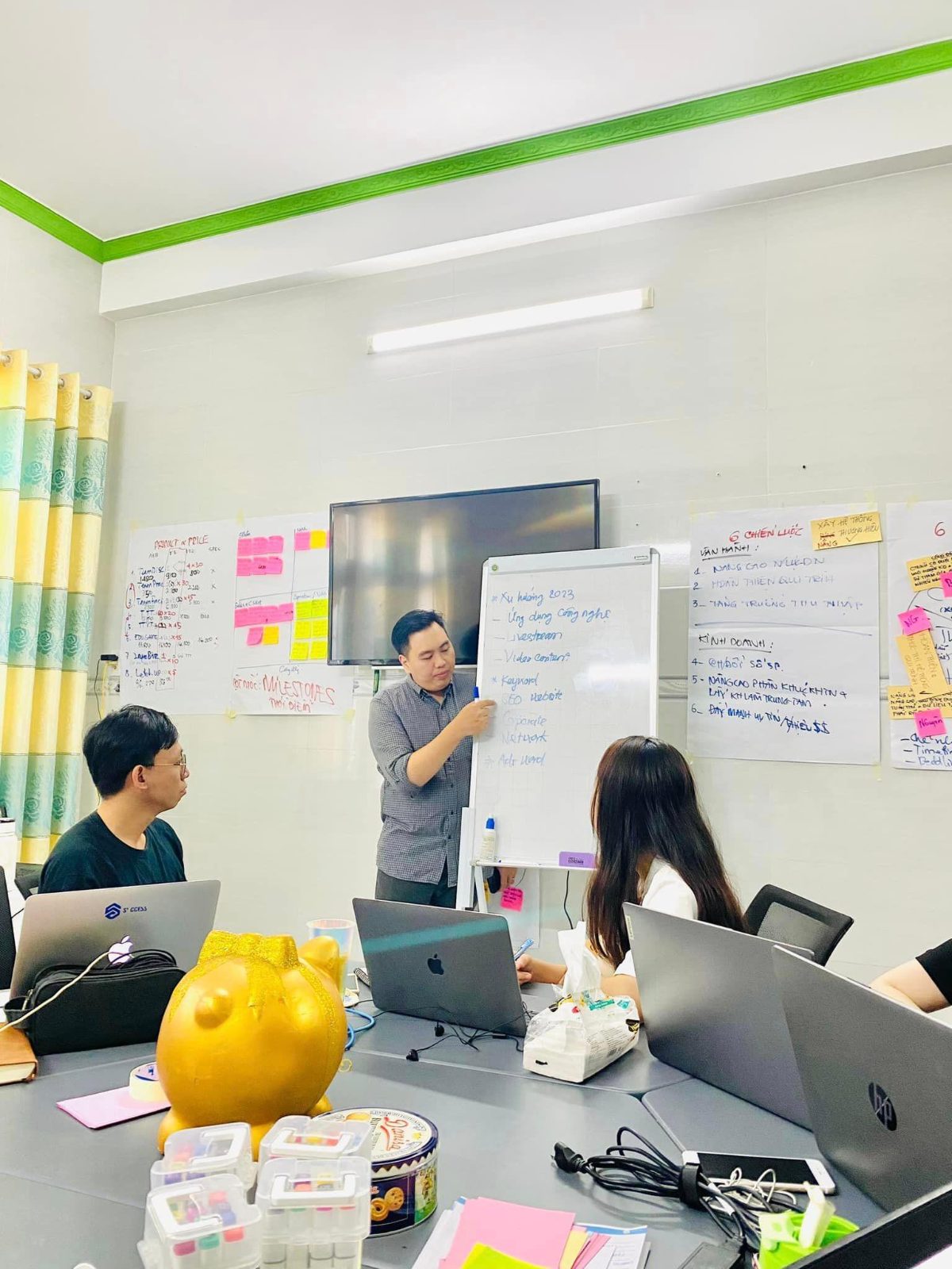 Anh Thanh đứng bên bảng trắng trình bày xu hướng marketing (công nghệ, livestream, video, SEO website) cho đồng nghiệp trong phòng họp, xung quanh dán kín kế hoạch và chiến lược